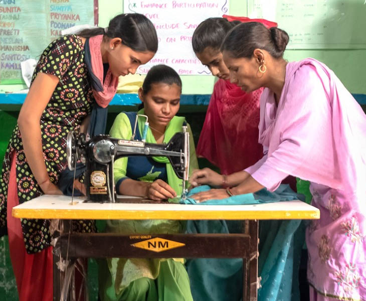 Three women stand with woman at sewing machine
